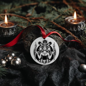 Könnte beinhalten: Ein weißer, runder Ornament mit einer Schwarz-Weiß-Illustration einer Frau mit drei Gesichtern, die Fackeln hält. Das Ornament hängt an einem roten Band. Der Hintergrund umfasst brennende Kerzen, silberne Kugeln und Tannenzweige.