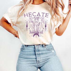Pu&ograve; includere: Una maglietta bianca con una grafica viola della dea greca Ecate, circondata da lune e stelle. Il testo "HEcate" &egrave; stampato in viola sopra la grafica.