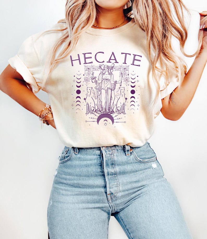 Pu&ograve; includere: Una maglietta bianca con una grafica viola della dea greca Ecate, circondata da lune e stelle. Il testo "HEcate" &egrave; stampato in viola sopra la grafica.