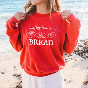 Può includere: Una felpa rossa con la scritta bianca "Loafing him was BREAD". Il design include tre illustrazioni di pane sopra la parola "BREAD". La felpa è di colore rosso pieno.