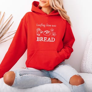 Può includere: Una felpa con cappuccio rossa con la scritta bianca "Loafing him was BREAD". Il design include illustrazioni di una sciarpa, pane e foglie. La felpa ha una tasca frontale e un cappuccio con coulisse.