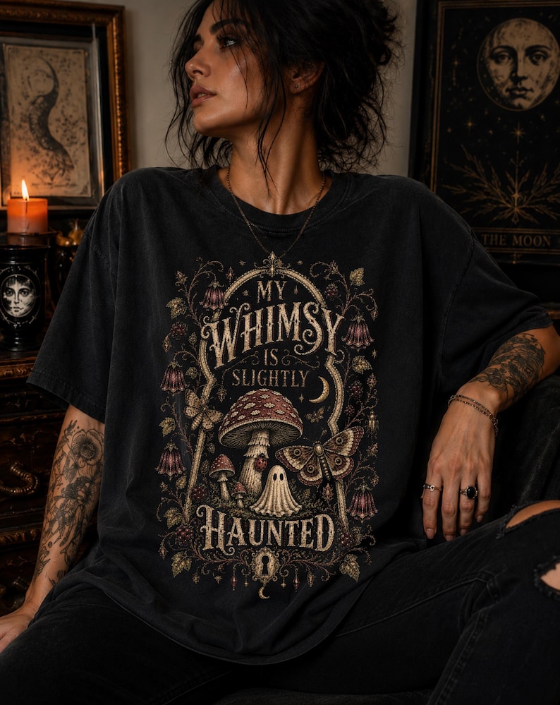 May include: A charcoal black t-shirt featuring a graphic print with the text "MY WHIMSY IS SLIGHTLY HAUNTED". The design includes a large mushroom, a small ghost figure, a moth, and intricate floral vines framed within an ornate archway.