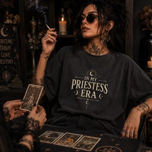 May include: A person wearing a dark gray t-shirt with the text "IN MY PRIESTESS ERA" in a white, stylized font. The person is wearing sunglasses and jewelry, and is holding a lit cigarette. Tarot cards are on the table.
