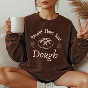 Può includere: Una felpa marrone con la scritta "Should Have Said Dough" in crema. Il design include un'illustrazione di una pagnotta. La persona tiene in mano una tazza bianca. Abbigliamento casual.