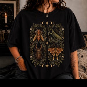 May include: A black short-sleeve t-shirt featuring a central graphic print of four insects: a bee, a praying mantis, a stag beetle, and a moth. The insects are framed by green foliage and small white flowers, accented with celestial star and moon symbols.
