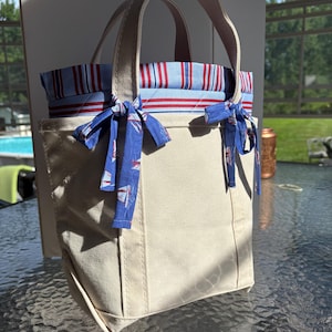May include: A beige canvas tote bag featuring two top handles and a blue fabric lining with red and white stripes. The exterior is accented with two blue fabric bows decorated with a sailboat pattern, tied around the base of the handles.
