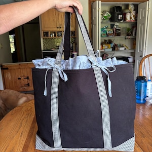 Può includere: Una borsa tote nera con un motivo a quadretti vichy bianchi e neri sulla base, sui manici verticali e sul bordo superiore. La borsa include una fodera interna grigio chiaro che sporge dal bordo superiore ed è chiusa da due piccoli lacci.