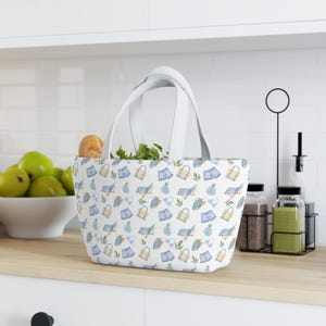 May include: A white tote bag featuring a repeating pattern of blue beach chairs, yellow tote bags, blue shorts, and green leaves. The bag has two white handles and a wide, rectangular base, showcasing a summer-themed design on a white fabric.