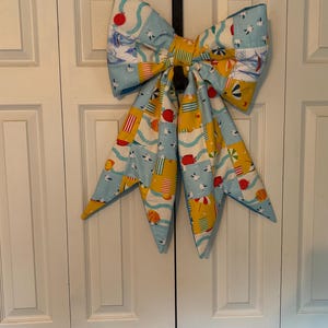 May include: A large decorative fabric bow featuring a repeating summer beach pattern. The design includes illustrations of seagulls, sailboats, red suns, yellow beach chairs, and striped umbrellas on a light blue and yellow background.