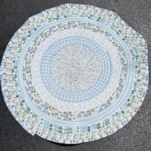 Puede incluir: Una alfombra redonda trenzada que presenta círculos concéntricos de tiras de tela. El diseño alterna entre patrones de cuadros azul claro y blanco, pequeños estampados florales azules y bandas de hojas verdes sobre un fondo blanco, cosidos.