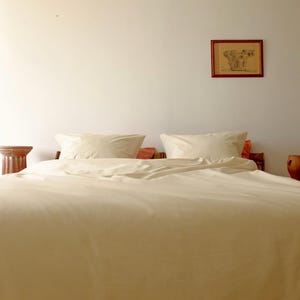 May include: A white bed with a white duvet cover and two white pillows. The bed is in a room with white walls and a small wooden table with a terracotta pot on it.