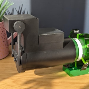 May include: A black telescope mount with a cylindrical tube and a green and white support structure. The mount is positioned on a wooden surface, with a pink pot of greenery in the background.
