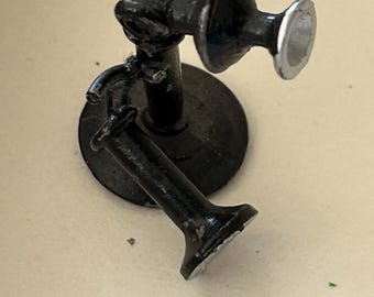 May include: A black, vintage-style telephone receiver with a circular base. The receiver has a curved handle and a flared mouthpiece. The earpiece has a silver rim. The telephone is made of metal and has a distressed finish.