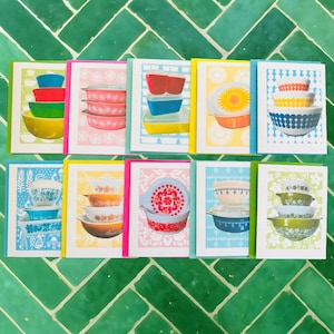 May include: A collection of greeting cards featuring colorful illustrations of vintage Pyrex bowls. Each card showcases a different arrangement of bowls in various colors, including red, blue, yellow, and green, set against patterned backgrounds. The cards are framed with bright borders.