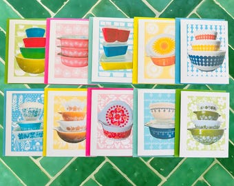 Könnte beinhalten: Eine Sammlung von Grußkarten mit farbenfrohen Illustrationen von Vintage-Pyrex-Schüsseln. Jede Karte zeigt eine andere Anordnung von Schüsseln in verschiedenen Farben, darunter Rot, Blau, Gelb und Grün, vor gemusterten Hintergründen. Die Karten sind mit hellen Rändern versehen.