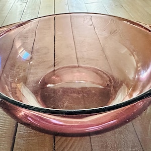 May include: A large, clear, pink glass mixing bowl with a black rim and two handles. The bowl is empty and sits on a wooden surface. The bowl is designed for mixing ingredients.