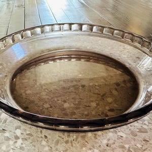 May include: A clear, round glass pie dish with a decorative rim and handles. The dish is transparent, allowing the surface beneath to be visible. The pie dish is designed for baking and serving pies.
