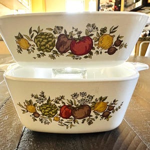 May include: Two white casserole dishes stacked on top of each other. Each dish features a colorful illustration of vegetables, including tomatoes, artichokes, and lemons. The dishes have handles on either side.