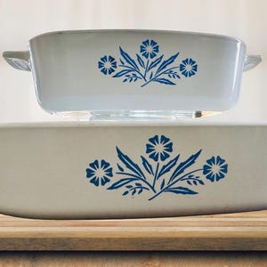 May include: Two white ceramic casserole dishes with blue floral designs. The larger dish is oval-shaped, and the smaller dish is rectangular with handles. Both are stacked on a wooden surface.