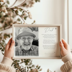 May include: A framed black and white photograph of a smiling man wearing a hat and glasses. The frame also includes a poem titled "Dad" with the dates 25.06.45-25.03.21. The frame is held by a person.