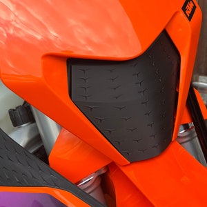 Könnte beinhalten: Nahaufnahme eines orangefarbenen Motorrad-Scheinwerfers mit einem schwarzen, strukturierten, rautenförmigen Gitter. Das Motorrad ist seitlich mit "KTM" beschriftet.