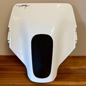 Op de afbeelding: Een witte motorfietskuip met een zwart middengedeelte. De kuip heeft een gebogen vorm met een handtekening in de linkerbovenhoek. Het object staat op een houten oppervlak.