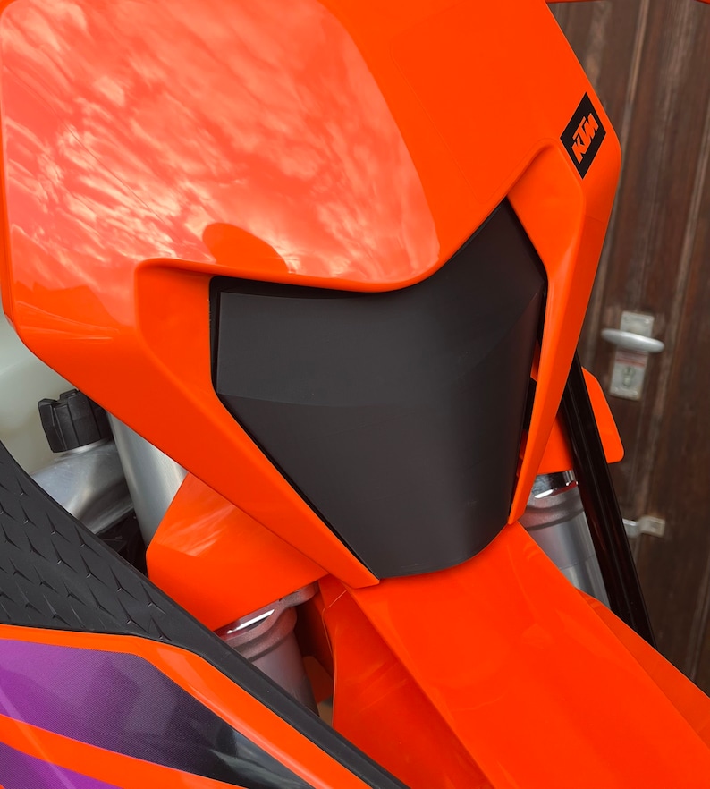 May include: Close-up of an orange motorcycle headlight with a black visor and the KTM logo on the side.