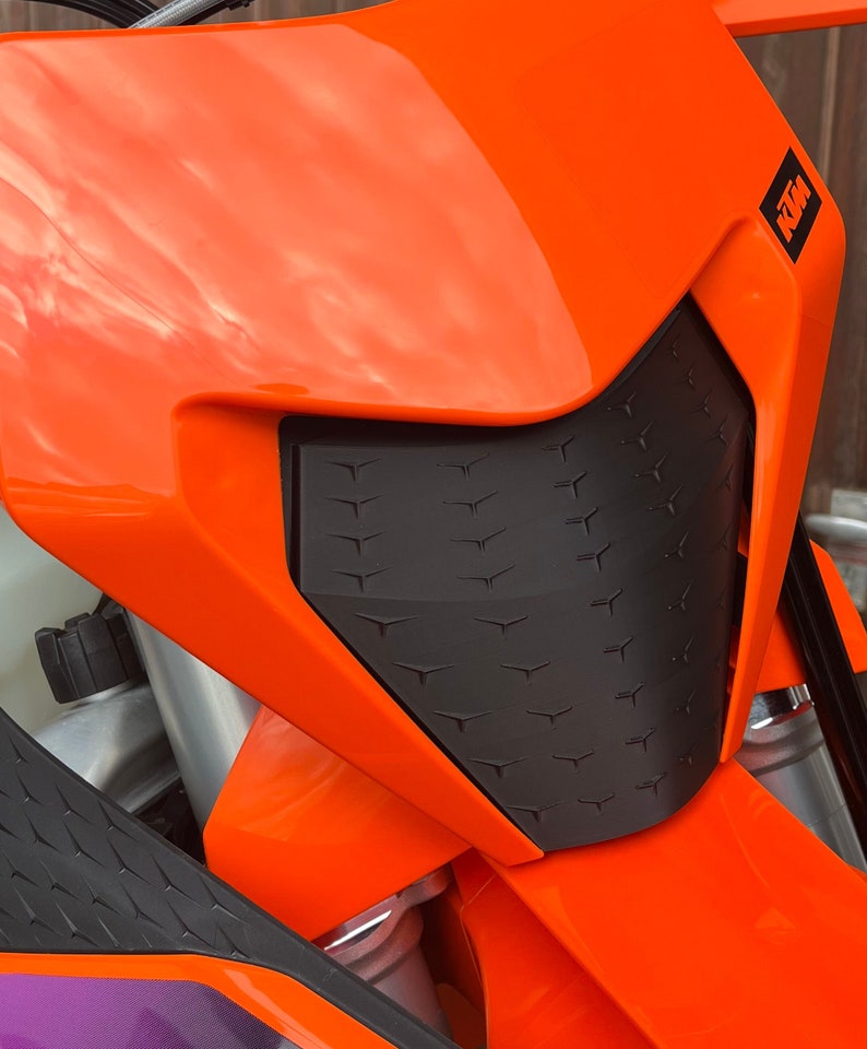 May include: Close-up of an orange motorcycle headlight with a black, textured, diamond-shaped grill. The motorcycle is labeled "KTM" on the side.