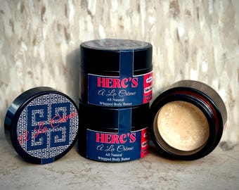 May include: Several black jars of Herc's À La Crème whipped body butter are displayed. One jar is open, revealing the product's light beige color. The labels feature red and blue accents with the product name and description.