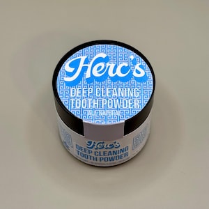 May include: A round black container of Herc's Deep Cleaning Tooth Powder. The lid features a blue and white design with the brand name in white script. The product name is in a bold, sans-serif font. The container is on a light gray surface.