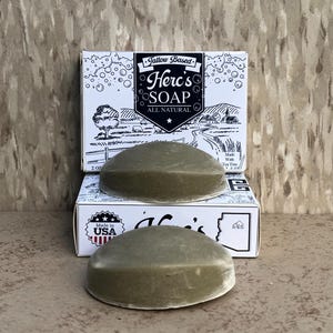 May include: Two oval-shaped, olive-green Herc's Soap bars sit in front of a white box with the Herc's Soap logo and the text "ALL NATURAL". The box also has a landscape illustration and the text "Made With Tea Tree".
