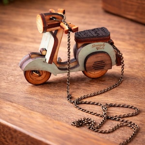 May include: Wooden scooter pendant necklace with a bronze chain. The scooter is light blue, brown, and tan with the words "Strangely Yours" on the back. The necklace is on a wooden surface.