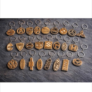 May include: A collection of wooden keychains featuring various car brand logos, including Nissan, Tesla, and Ford. Each keychain has a silver ring and is made of light brown wood. The keychains are arranged on a dark gray surface.
