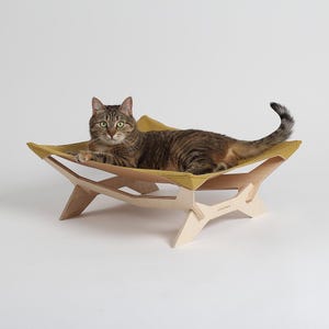 May include: A tabby cat lounges on a modern cat bed. The bed features a mustard-yellow fabric hammock suspended by a light-colored wooden frame. The cat is brown and tan with green eyes.