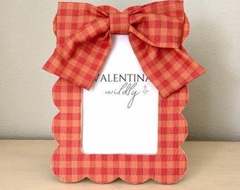 May include: A rectangular picture frame with a scalloped edge, covered in an orange and red gingham fabric. A matching fabric bow is attached to the top center. The white insert inside the frame features the text VALENTINA and the word wildly.