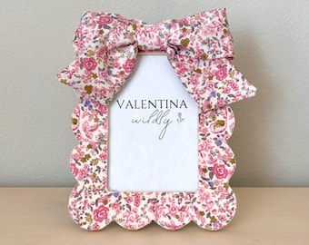 May include: A white picture frame with a scalloped edge and a floral pattern. The frame has a pink bow on top and the text "VALENTINA wildly" inside.