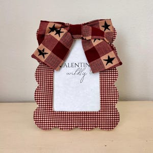 Op de afbeelding: Decoratieve fotolijst met een geschulpte rand, bedekt met een rood-wit geruit patroon. Een grote strik met zwarte steraccenten zit bovenop de lijst. De tekst "VALENTINE wildly" is zichtbaar.