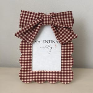 Könnte beinhalten: Ein dekorativer Bilderrahmen mit einem gewellten Rand und einer großen Schleife oben. Der Rahmen und die Schleife weisen ein rot-weißes Vichy-Muster auf. Der Text "VALENTINA wildly" ist im Rahmen sichtbar.