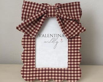 May include: A decorative photo frame with a scalloped edge and a large bow at the top. The frame and bow feature a red and cream gingham pattern. The text "VALENTINA wildly" is visible within the frame.