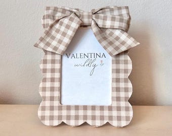 May include: A decorative photo frame with a scalloped edge and a large bow at the top. The frame and bow feature a tan and white gingham pattern. The text "VALENTINA wildly" is printed on the white space inside the frame.