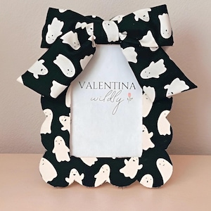 Op de afbeelding: Decoratieve fotolijst met een zwarte achtergrond en een wit spookpatroon. De lijst heeft een geschulpte rand en een grote strik aan de bovenkant. De tekst "VALENTINA wildly" is in het midden zichtbaar.