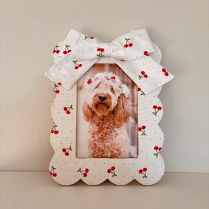 Op de afbeelding: Wit fotolijstje met een gegolfde rand en een witte strik met rode kersenprint. Het lijstje bevat een foto van een hond die een bloemenkrans draagt.