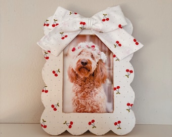 May include: White picture frame with a scalloped edge and a white bow with red cherry print. The frame has a photo of a dog wearing a flower crown.
