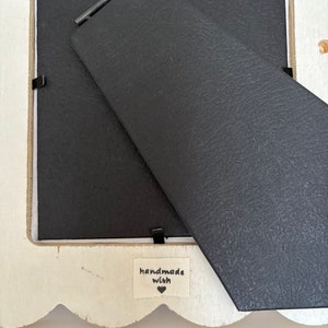 May include: Back of a white scalloped-edge picture frame with a black backing and stand. A small fabric tag reads "handmade with" and a heart symbol.