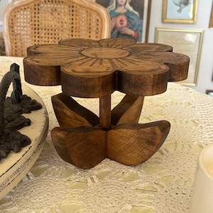 May include: A small, brown, wooden pedestal stand shaped like a flower. The top platform is circular with carved petal edges and a central sunburst design. It is supported by a central pillar and a base composed of four curved wooden petal shapes.