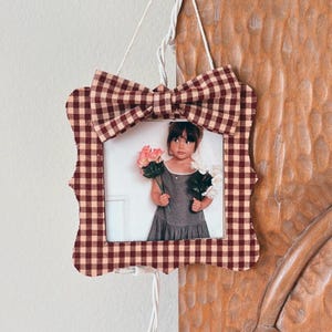 May include: A decorative photo frame with a gingham pattern in burgundy and cream, featuring a bow at the top. The frame holds a photograph of a young girl holding flowers. The frame is hanging on a wooden surface.