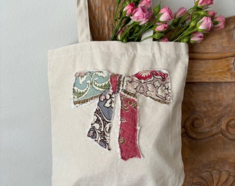 May include: A beige canvas tote bag with a large fabric bow made from patchwork fabric in shades of blue, green, pink, and brown. The bow has a frayed edge.