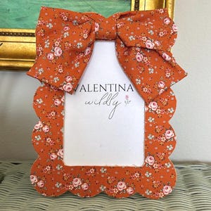 May include: A scalloped picture frame covered in burnt orange fabric with a floral pattern of pink roses and small white flowers. A large fabric bow sits at the top. The frame contains a white insert with the text VALENTINA wildly and a small rose.