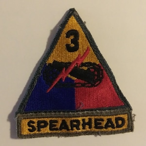 May include: A triangular patch with a yellow background, a blue and red section, and a black and red symbol. The number "3" is in the center of the yellow section. The text "SPEARHEAD" is on the bottom of the patch.