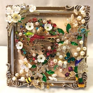 Pode incluir: Uma moldura quadrada prateada e ornamentada contendo uma colagem de joias vintage. O arranjo apresenta um pássaro vermelho e verde, um abelhão, um beija-flor, detalhes florais brancos, pérolas e strass coloridos sobre um fundo claro.